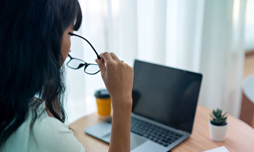 An Asian woman, wearing glasses, sits focused on her laptop in a bright, well-lit room. As a freelancer, she knows how crucial it is to protect her eyes from strain during long hours of screen time. By developing sustainable habits, like adjusting screen brightness, taking regular breaks, and practicing eye exercises, she ensures her vision stays sharp and her workday stays comfortable.