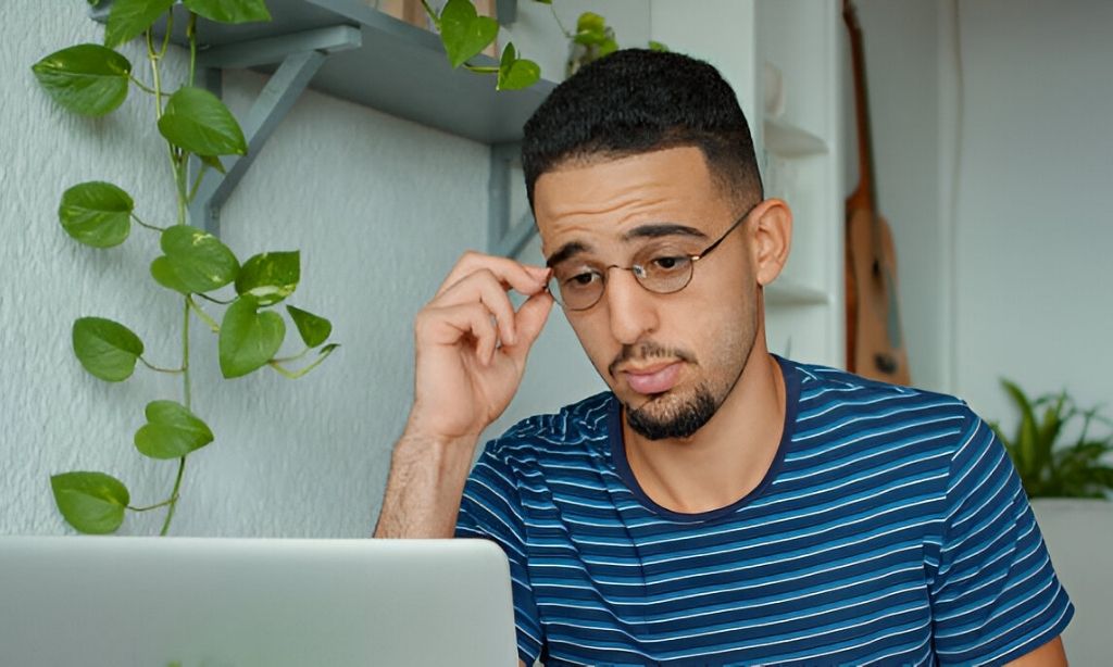 A young man looks frustrated, rubbing his eyes after a long session in front of his laptop. As a freelancer, he’s become all too familiar with the effects of prolonged screen exposure. Now, he’s taking proactive steps to manage his eye strain—reworking his workstation setup, incorporating frequent eye breaks, and learning strategies to prevent discomfort and maintain productivity.